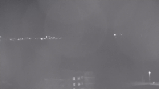 A monochrome nighttime view showing faint lights in the distance, possibly from buildings or streetlights. The foreground is dark and obscured, with a vague outline of structures. The scene has a hazy, indistinct quality, making it difficult to discern specific features or details.
