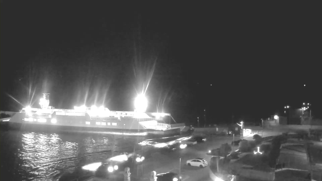 A brightly lit ferry is docked at a harbor during the night. The water reflects the lights from the ferry, creating shimmering patterns. Nearby, several vehicles are parked along the waterfront, with headlights visible. The surrounding area is dark, with a few distant lights indicating buildings or structures.