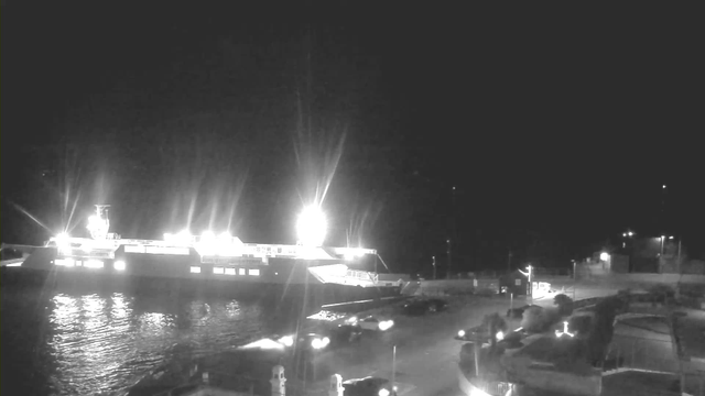A brightly lit ferry is docked at night, illuminated by strong lights on its upper deck, reflecting on the water. The foreground shows a dark harbor area with parked vehicles, and a few scattered light sources, indicating structures nearby. The scene is predominantly black and white, conveying a high-contrast nighttime atmosphere.