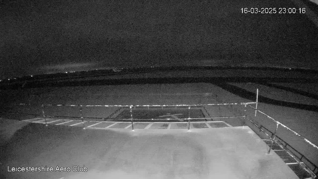 A dimly lit outdoor scene captured by a webcam at the Leicestershire Aero Club. The foreground features a flat surface, possibly an airport runway, with some lines and markings visible. A railing, consisting of vertical posts and horizontal bars, runs along the edge of the surface. In the distant background, a dark sky is present, with faint lights suggesting either cities or vehicles far away. The scene appears quiet and still, with no discernible activity. The date and time displayed in the corner indicate 16th March 2025 at 11:00 PM.