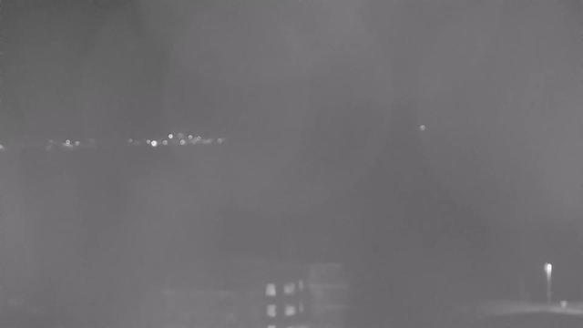A low-light image captured from a webcam, showing a dark landscape with faintly illuminated spots resembling distant city lights on the horizon. The foreground features a blurred structure, possibly a building, with some lit windows. The overall scene has a gray tone, emphasizing the night setting.