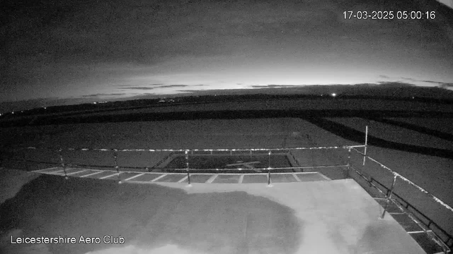 A dimly lit scene showing an outdoor area at dawn. In the foreground, there is a flat surface with a faint outline of a helicopter landing pad marked by a cross. There are railings along one side, creating a barrier. The sky is mostly cloudy with dark blues and grays, hinting at the early morning light on the horizon. In the distance, a few lights can be seen twinkling faintly. The bottom left corner has the text "Leicestershire Aero Club."