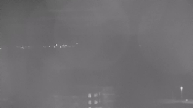 A low-contrast black and white webcam image showing a blurred view of lights in the distance. The image features some faint urban or natural landscape outlines, but details are not clear, making it difficult to discern specific objects or features. The background is dark, with small, bright spots representing lights possibly from buildings or streetlights, and a hint of a structure in the lower right corner.