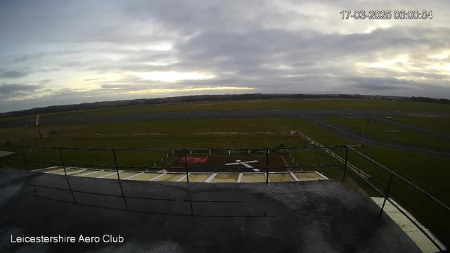 A view from a webcam located at the Leicestershire Aero Club, capturing an expansive landscape at dawn. The foreground features a flat rooftop structure with a railing and sections of visible roofing. Below, an airstrip extends into the distance, bordered by grassy fields. A white cross is marked on the ground, and there are scattered white posts along the edges of the airstrip. The sky above is overcast, with clouds creating a soft, muted light, suggesting early morning. The timestamp at the top right indicates the image was taken on March 17, 2025, at 6:00 AM.