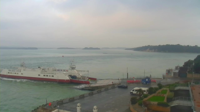 A ferry is docking at a harbor with a gray sky above. The water is calm and slick, reflecting light as the ferry approaches the shore. In the background, green hills rise along the coastline. Nearby, vehicles are parked on a paved area, with some bushes and a few planters visible. The atmosphere appears overcast and tranquil, indicative of a cloudy day.