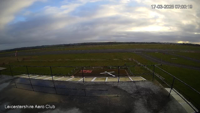 A view from a webcam at the Leicestershire Aero Club, showing an expansive airfield. The scene captures a cloudy sky with patches of blue, and a grassy landscape stretching out to the horizon. A large rectangular area marked with a white cross is visible on the ground below. There are wooden fences surrounding sections of the airfield and a windsock indicating wind direction. In the distance, an airport runway is visible, along with some scattered markings on the ground. The image is set during early morning, as indicated by the timestamp.