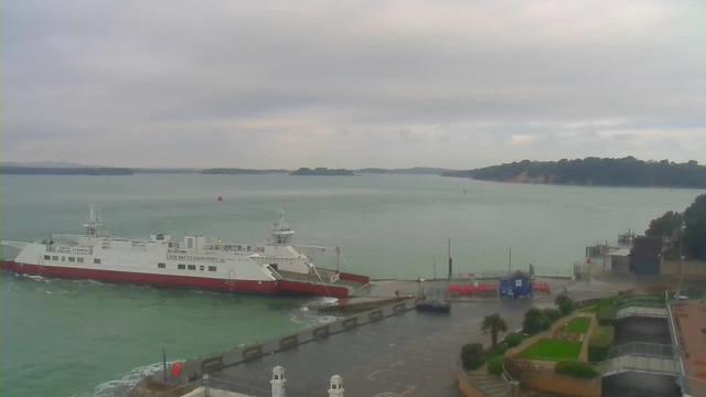 A ferry boat is docked at a pier beside a calm, greenish-blue body of water. The sky is overcast with gray clouds, and the distant shore is visible in the background, with trees and a small hill. The pier has a few structures, and there are some people and vehicles near the waterfront. Green grass and landscaped areas are visible on the right side of the image.