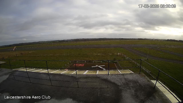 A view from a webcam at Leicestershire Aero Club, showing a vast open area with a grassy field and a runway in the background. The sky is overcast with gray clouds, and scattered light patches are visible. In the foreground, there is a railing and a paved surface leading to a grassy area with a white ‘X’ marked, likely indicating a landing zone. Fencing is visible around the grassy area, and there are signs present but not clear from the image. The image is timestamped and dated.