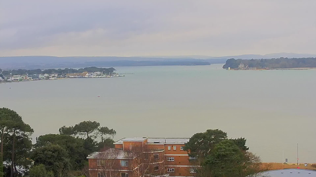 A view overlooking a calm body of water with a hazy sky above. On the left, a marina features various boats and buildings along the shoreline. In the background, gentle hills and trees line the opposite side of the water. In the foreground, there are some trees and a building with orange brick exteriors. The overall atmosphere is serene, with muted colors and an overcast sky.
