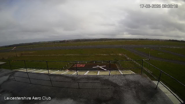 A view from a webcam at Leicestershire Aero Club showing a grass airfield under a cloudy sky. In the foreground, there is a balcony railing, and below, a grassy area with a red and white helipad marking. The runway extends into the distance, lined with white fences and a few scattered patches of grass. The date and time are displayed in the top right corner.