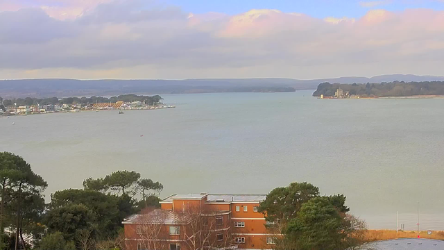 A wide view of a lake or estuary under partly cloudy skies. In the foreground, there are trees and a building with orange walls and a flat roof. The water is calm and reflects the soft colors of the clouds. On the opposite shore, there are several buildings and a small boat, while further along is a small marina with boats docked. In the distance, rolling hills and a wooded area can be seen, indicating a natural landscape.