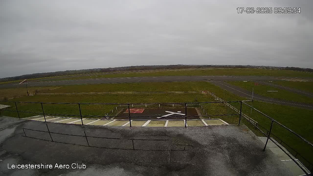 A wide view of a grassy airfield under a cloudy sky, with a runway visible extending into the distance. In the foreground, there are railings, suggesting the camera is positioned on a balcony or upper level. A large white cross is painted on the ground near the edge of the field, while red markings can be seen in the grass. The scene is calm and devoid of any aircraft or people. The image has a timestamp in the top corner displaying the date and time.
