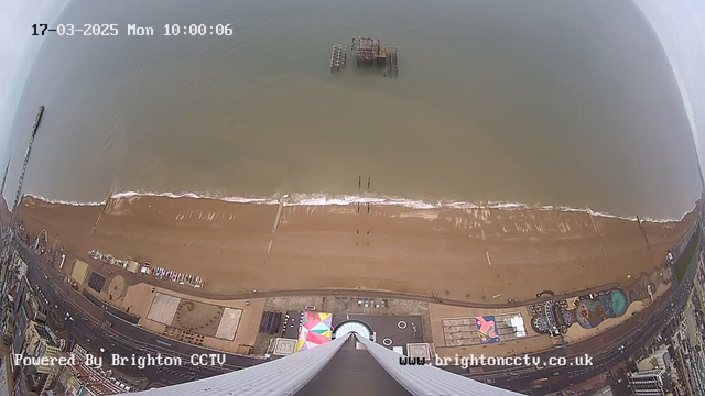 A coastal view captured from a high angle, showing a sandy beach bordered by the ocean. On the beach, there are a few people visible along the shore. There is an abandoned pier structure partially submerged in the water. Below the camera, a colorful amusement park area is seen, along with a circular building and several scattered structures. The water appears calm with gentle waves. The sky is mostly overcast. The date and time are displayed in the top left corner.
