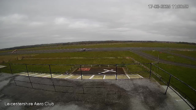 A cloudy day at a small airfield. In the foreground, a wooden railing is visible along a balcony with a concrete surface. Below, there is a grassy area with a red square and a white cross painted on it. A light aircraft is positioned on the runway in the midground, with taxiways visible in the surrounding area. The sky appears overcast, giving a muted ambiance to the scene. In the background, there are trees and additional fields stretching out, indicating a rural setting.