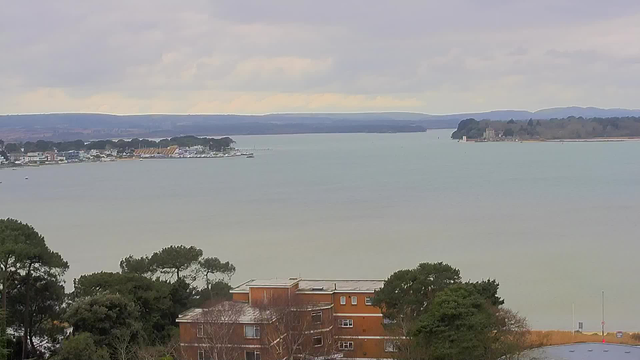 A view of a calm body of water with a distant shoreline lined with trees and buildings. In the foreground, there is a large brown building partially obscured by greenery. The sky is overcast with soft grey clouds, and the horizon features rolling hills. Small boats can be seen near the shore and further out on the water.
