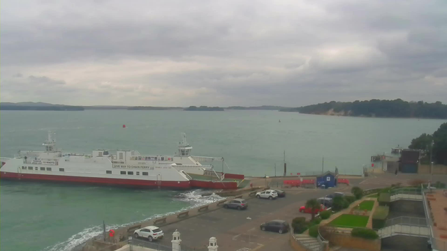 A view of a harbor under a cloudy sky. In the foreground, a large white and red ferry is docked, showing a sign that reads "GIVE WAY TO CHAIN FERRY." Several parked cars are visible along the shoreline, and there are some trees and a slope leading down to the water. The water appears choppy with small waves, and there is an island in the distance with a rocky coast. The overall atmosphere is calm, with muted colors from the clouds and sea.