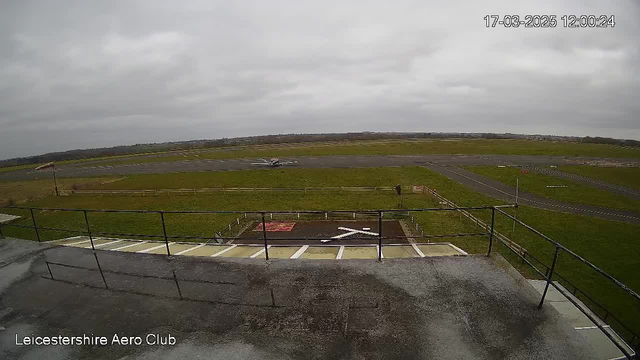 A cloudy sky is visible above a grassy airfield. A small aircraft is positioned on the runway to the left, while a person appears to be walking in the foreground. A flat rooftop railing surrounds the view, with white markings on the ground suggesting a landing area. The scene conveys a calm atmosphere typical of an aerodrome.
