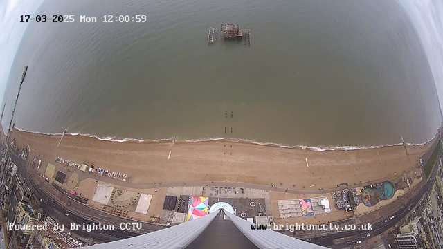Aerial view of a beach scene on a cloudy day. The shoreline features a wide expanse of tan sand bordered by calm water with small waves. There is a dilapidated pier extending into the water, and several people are seen walking along the beach. In the lower part of the image, there are structures, including a colorful area with distinct patterns, likely for amusement. The scene appears to be taken from a tall building or structure, looking down towards the coastline.