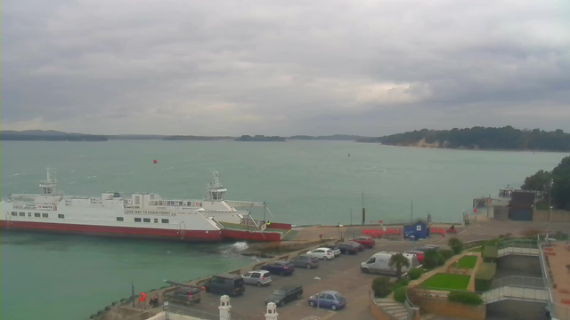 A ferry is docked at a pier on a cloudy day, with the water appearing choppy. Several cars are parked along the waterfront, and there are green areas with low plants nearby. In the background, a wooded shoreline stretches along the horizon, with a few gentle hills. The sky is overcast, indicating the possibility of rain.