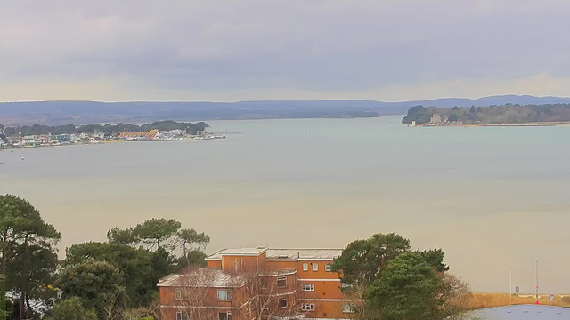 A view of a calm body of water under a cloudy sky, with distant hills in the background. In the foreground, there are trees and a building with orange walls. Along the shoreline, various boats are visible, and there are several houses and structures lining the water’s edge. The overall atmosphere is serene and slightly overcast.