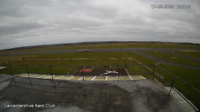 A view from a webcam showing a partially cloudy sky above an airfield. In the foreground, there is a railing and a flat rooftop surface. Below, the airfield is visible with an aircraft on the runway in the distance. The grass surrounding the runway is green, and there are structures and markings on the ground, including a red area and a white cross shape. The scene suggests it's midday, and the ambiance appears calm.