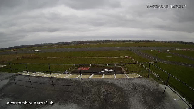 A view from a webcam at an airport featuring a gray, overcast sky. In the foreground, there is a flat surface with a railing, and a grassy area extends into the distance. A runway is visible to the right, with a small aircraft parked on it. The scene is serene, with a few scattered trees and fields beyond the airport area. The image timestamp indicates it is 2:00 PM on March 17, 2025.