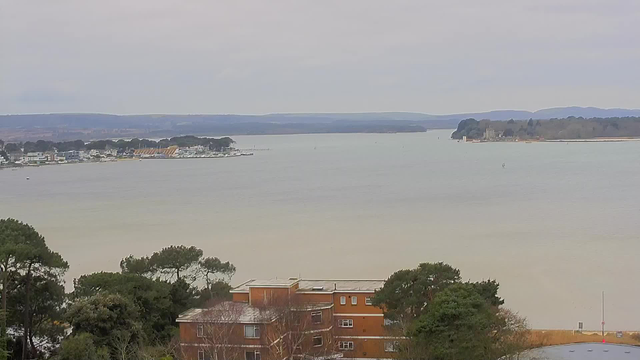 A wide view of a calm body of water, with a shoreline on the left featuring various buildings and boats. In the foreground, there are some trees and an orange building partially visible. The scene is set against a cloudy sky and distant hills are visible in the background, creating a serene atmosphere. The water has a light brown hue and appears still.
