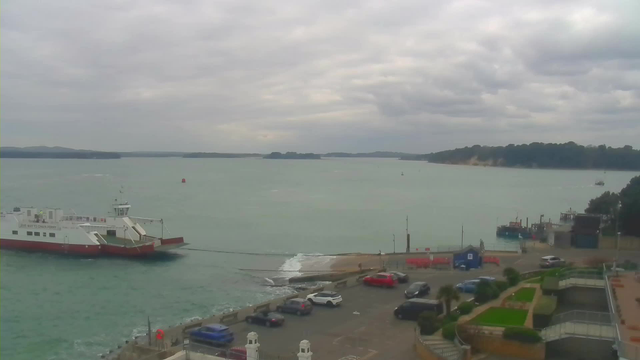 A view of a calm harbor under a cloudy sky. In the foreground, there is a parking lot filled with cars, including red, blue, and silver vehicles. A large white ferry with red accents is docked near the water's edge, with its ramp extended. To the right, there is a small dock with a couple of boats visible and a background of green hills in the distance. The water appears a muted greenish-blue. Closer to the shoreline, there are sandy areas where waves gently lap at the shore.