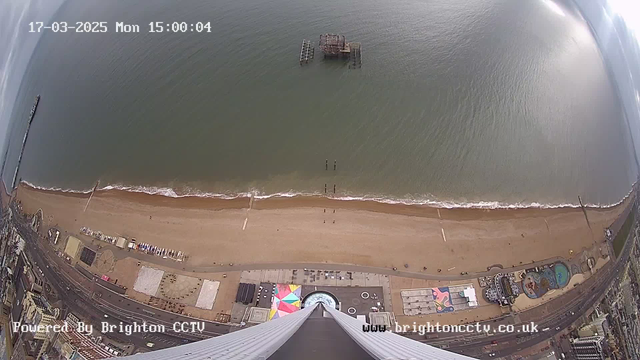 Aerial view of a sandy beach next to the ocean on a cloudy day. The shoreline has gentle waves lapping at the sand. A partially submerged pier can be seen in the water, along with a few vertical posts. Near the bottom of the image, colorful beach huts and structures are visible, along with a circular structure. The scene captures a mixture of sand and water, with no people prominently visible. The image features a timestamp indicating the date and time of the capture.