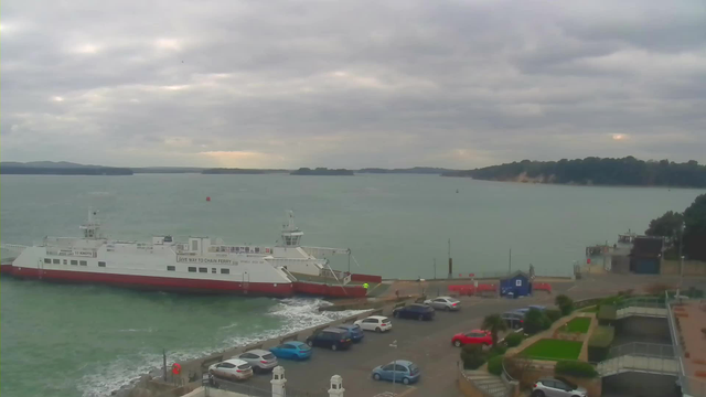 A view of a harbor with a large white and red ferry docked at the edge of the water. The ferry is stationary and has text indicating its purpose. The water is choppy, with visible waves. In the foreground, there is a parking area filled with various cars in different colors. Beyond the ferry, the coastline features green hills or trees, and the sky is overcast with gray clouds. The scene suggests a cool, possibly windy day.