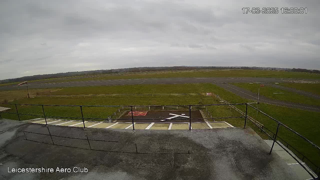 A view from an elevated position at the Leicestershire Aero Club. The foreground shows a flat surface with a white cross marking, surrounded by green grass and a fence. In the background, there is a long gray runway stretching to the horizon, with patches of grass visible alongside. The sky is overcast with gray clouds, giving a soft lighting to the scene.