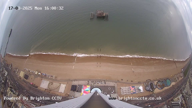 Aerial view of a beach with golden sand and gentle waves lapping at the shore. In the foreground, a wide, surfacing area is visible, with a central structure that has a circular opening. There are some beach amenities and colorful canopies on the sand. In the distance, an old, abandoned pier stretches out into the sea. The sky above is partly cloudy, with soft light illuminating the scene. The image also displays a timestamp indicating March 17, 2025, at 16:00:32.
