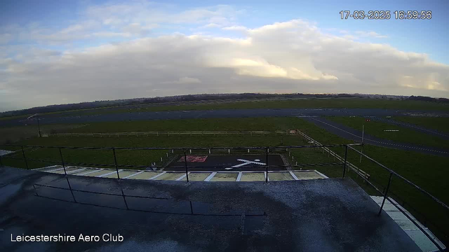 A view from a webcam showing a wide, open landscape with a runway at the Leicestershire Aero Club. The foreground features a railing and a section of the ground with a red square and a white cross marking. In the background, there is a grassy area and a runway extending into the distance, under a partly cloudy sky.