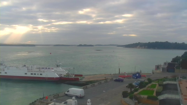 A coastal scene features a calm harbor with a ferry docked on the left side. The ferry is white with a red stripe along the bottom and has visible signage. The water is a soft blue-green, with gentle ripples reflecting a cloudy sky. Sunlight beams through breaks in the clouds, illuminating the horizon where dark silhouettes of hills can be seen in the distance. On the shore, vehicles, including a white van and a blue car, are parked near a landscaped area with plants. The atmosphere is tranquil and overcast, conveying a soothing coastal environment.
