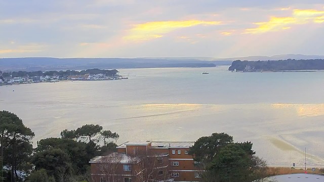 A landscape view of a calm body of water extends towards the horizon, with gentle waves reflecting a soft golden light from a cloudy sky. In the foreground, there are trees and buildings, while the middle ground features a marina with boats moored. In the distance, hills fade into the background under overcast skies. A small vessel is visible moving in the water.