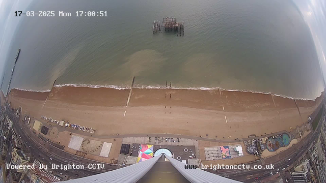 An aerial view of a beach and shoreline. The image shows a sandy beach with a few people walking along it and a pier extending into the ocean. In the water, just off the shore, there is an old, partially submerged pier structure. The beach is lined with colorful amusement equipment and structures, with the ocean appearing calm under a slightly cloudy sky. The timestamp in the top left corner indicates the date and time.