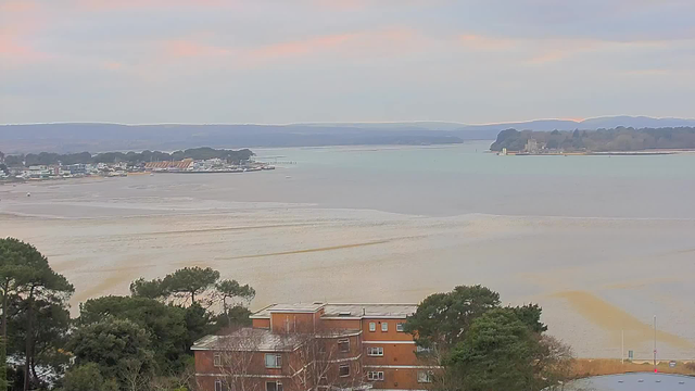 A panoramic view of a calm body of water with gentle waves, bordered by low hills in the background. On the left, a small marina with boats is visible, while various buildings, including some low-rise apartments, are situated nearer to the foreground. The sky is overcast with soft pastel colors suggesting early morning or dusk light. The shoreline features patches of sand and water, creating a serene landscape.