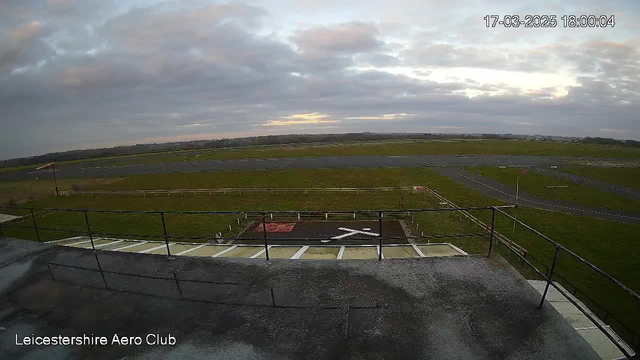 A view from the Leicestershire Aero Club webcam shows a grassy airfield under a cloudy sky during twilight. In the foreground, there is a flat rooftop with a railing. Below the rooftop, a red square with a white 'X' is marked on the ground, indicating a designated landing area. The airfield features a long, paved runway extending into the distance, flanked by low grass on both sides. There are various light poles and signs scattered around the area, and the horizon in the background is obscured by clouds.