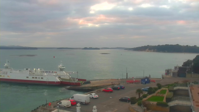 A view of a calm harbor with a ferry docked on the left. The ferry is white with red accents, and there are vehicles, including red and white cars and a van, parked nearby on the concrete dock. In the background, the water is a soft blue-gray, with small islands visible in the distance. The sky is overcast with hints of pink and orange, indicating sunset. To the right, there are buildings and greenery along the shore.