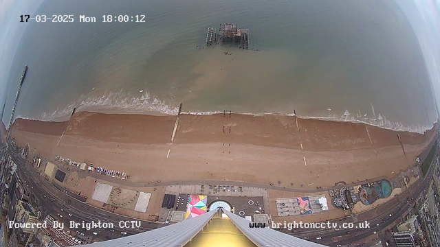 Aerial view of Brighton beach taken from a high vantage point. The image shows a sandy shoreline with waves gently lapping at the beach. A pier with a partially submerged structure is visible in the water. On the beach, a few people can be seen walking near the water's edge. In the foreground, a colorful patterned area is discernible, along with buildings and a pool area. The sky is predominantly gray with minimal cloud coverage. The timestamp indicates the image was taken on March 17, 2025, at 18:00.