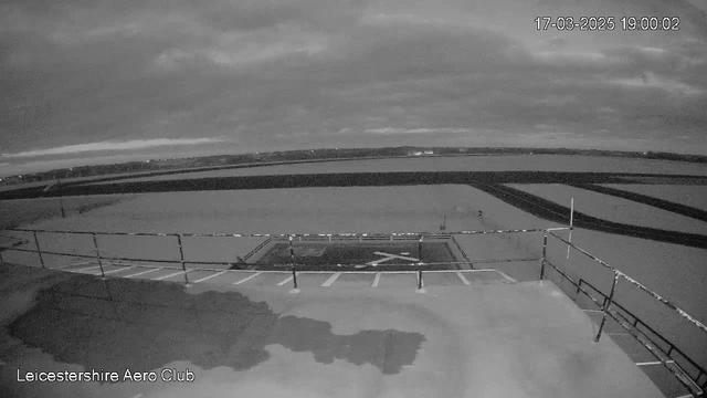 A black and white image from a webcam showing a flat rooftop with a railing in the foreground. In the center, there is a marked helipad with an "X" symbol. The background features a vast open area of land and water, with cloudy skies above. The horizon is slightly visible in the distance, indicating a rural or semi-urban setting. A timestamp in the corner reads "17-03-2025 19:00:02," and the text "Leicestershire Aero Club" is displayed at the bottom.