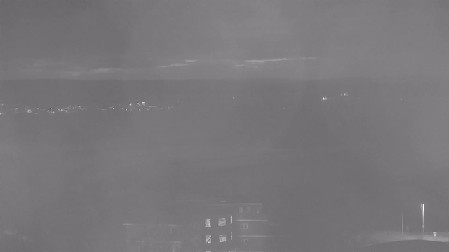 A dimly lit landscape at dusk, with faint outlines of distant hills and a cloudy sky. In the foreground, blurred shapes suggest a building with a few lit windows. There are small, twinkling lights that may represent a town or street below. The scene has a monochromatic, grayscale tone, creating a tranquil yet moody atmosphere.