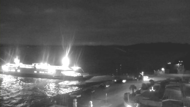 A dark waterfront scene at night featuring a brightly lit ferry docked near the shore. The water reflects the lights, creating a shimmering effect. The background includes distant hills and some faint lighting from vehicles and structures along the waterfront. The image conveys a serene nighttime atmosphere.