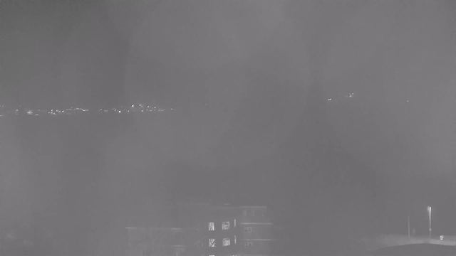 A dimly lit night scene captured by a webcam. In the foreground, there is a dark building with a few illuminated windows. In the background, a faint outline of a landscape can be seen, with scattered lights possibly indicating distant buildings or cars. The image appears hazy, with a grayish tint, reducing visibility and clarity.
