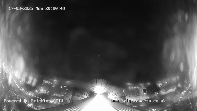 A dark, blurred image captured from a webcam, showing a faintly illuminated area at the center, likely the view from a tall building. The edges have a soft focus with some light reflections or rain droplets, while the corners display indistinct urban elements. The timestamp at the top indicates "17-03-2025 Mon 20:00:49" and branding at the bottom reads "Powered By Brighton CCTV" along with a website link.