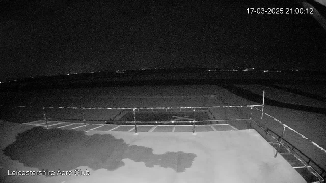 A dark night scene captured by a webcam showing a flat rooftop area with a railing. The surface appears wet, with a shadowy puddle reflecting some light. In the background, faint lights from distant sources are visible, indicating a landscape beyond the rooftop. The image is devoid of any prominent features, focusing mainly on the roof and the distant glow of lights.
