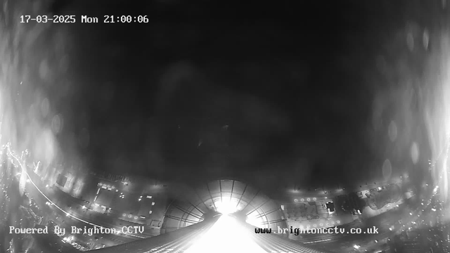 A black and white image from a webcam showing a cityscape at night. The scene is mostly dark, with some areas illuminated by streetlights and vehicle headlights, creating streaks of light. The date and time are shown in the upper left corner, indicating March 17, 2025, at 21:00:06. The bottom of the image has text that reads "Powered By Brighton CCTV" and a website URL.