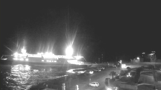 A brightly lit ferry docked at night, reflecting light off the calm water. The surrounding area features a pathway leading to the ferry, with several parked cars and faint lights in the background indicating nearby structures. The scene is primarily dark with strong highlights from the ferry's lights.