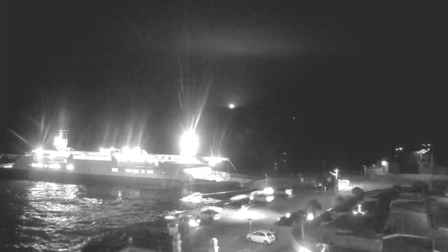 A brightly lit ferry is docked at the edge of the water during nighttime. The vessel has multiple lights illuminating its deck and the surrounding area, creating reflections on the water's surface. In the foreground, a road curves around the dock area, lined with a few parked and moving vehicles. The sky is dark, with some distant light visible above the horizon.