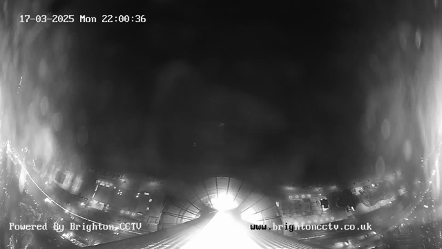 A black and white image captured from a webcam shows a view of a street at night. The center of the image is bright, likely from streetlights, while the edges appear darker. In the lower part of the image, faint outlines of buildings and vehicles can be seen. The timestamp in the top left corner indicates the date and time as March 17, 2025, at 10:00:36 PM. The bottom left corner displays "Powered By Brighton CCTV," and the bottom right corner features the website address "www.brightoncctv.co.uk."